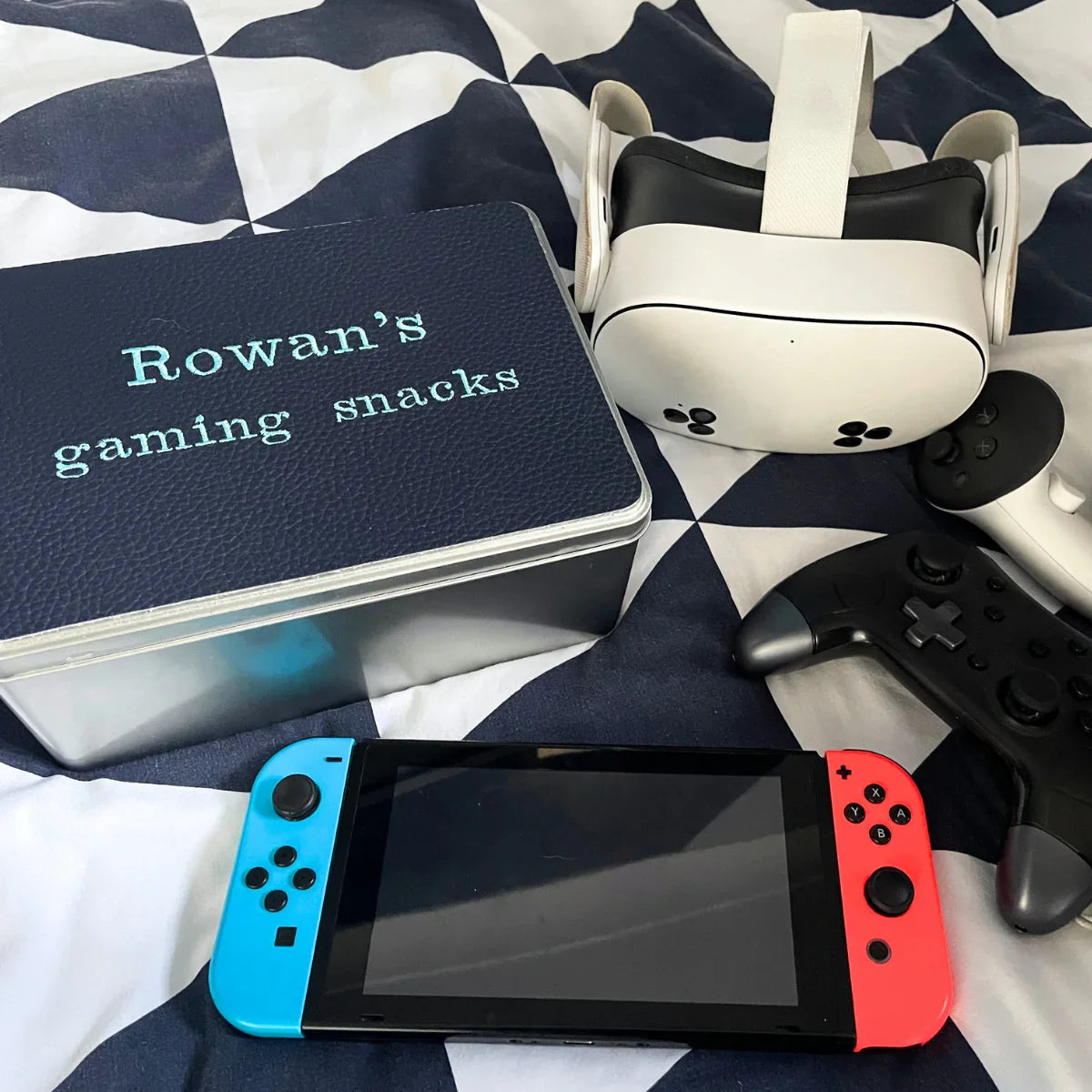 Gaming snacks tin with Nintendo Switch, controllers, and VR headset on patterned bedding