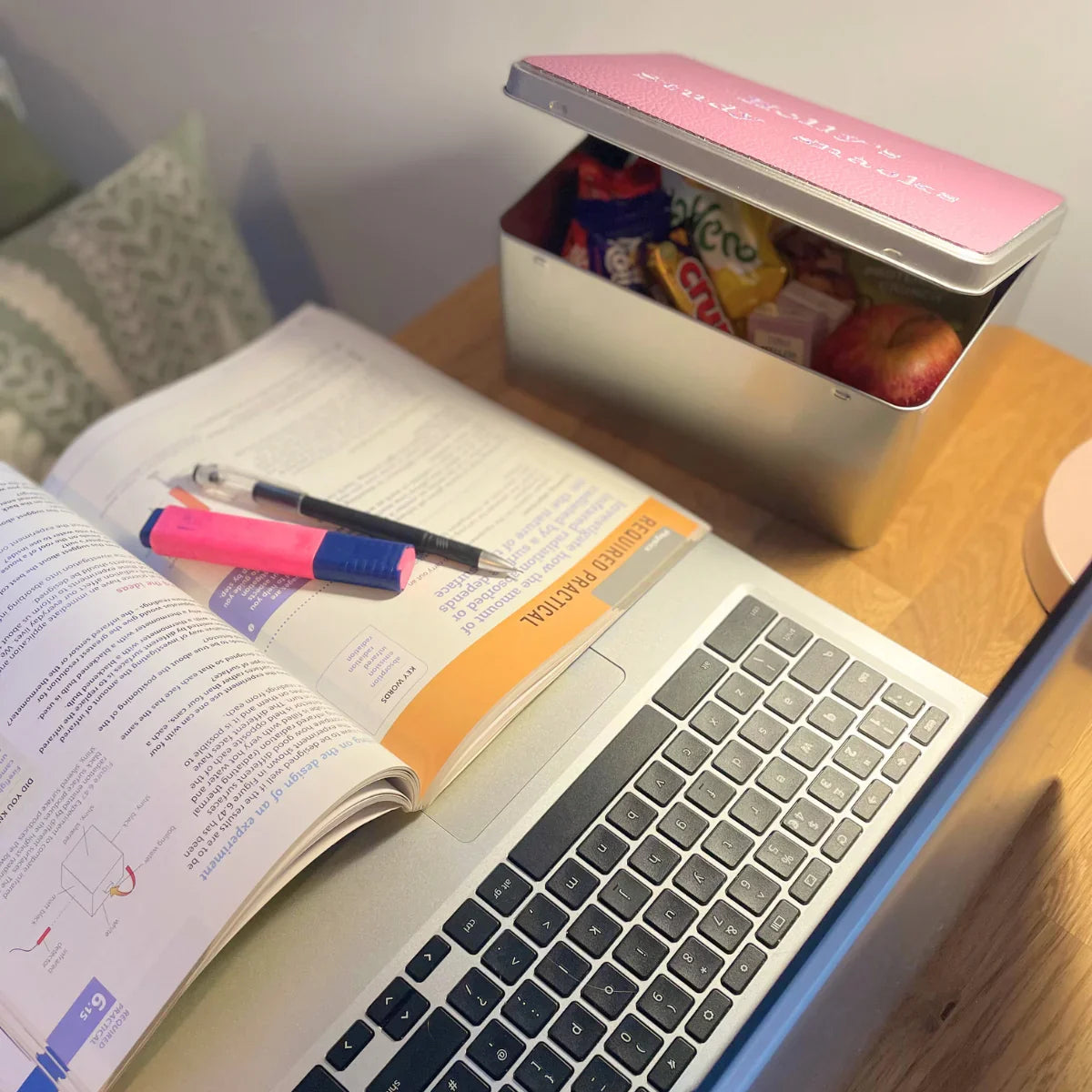 Study snacks box with fruit and treats on desk beside laptop, open textbook, and pens